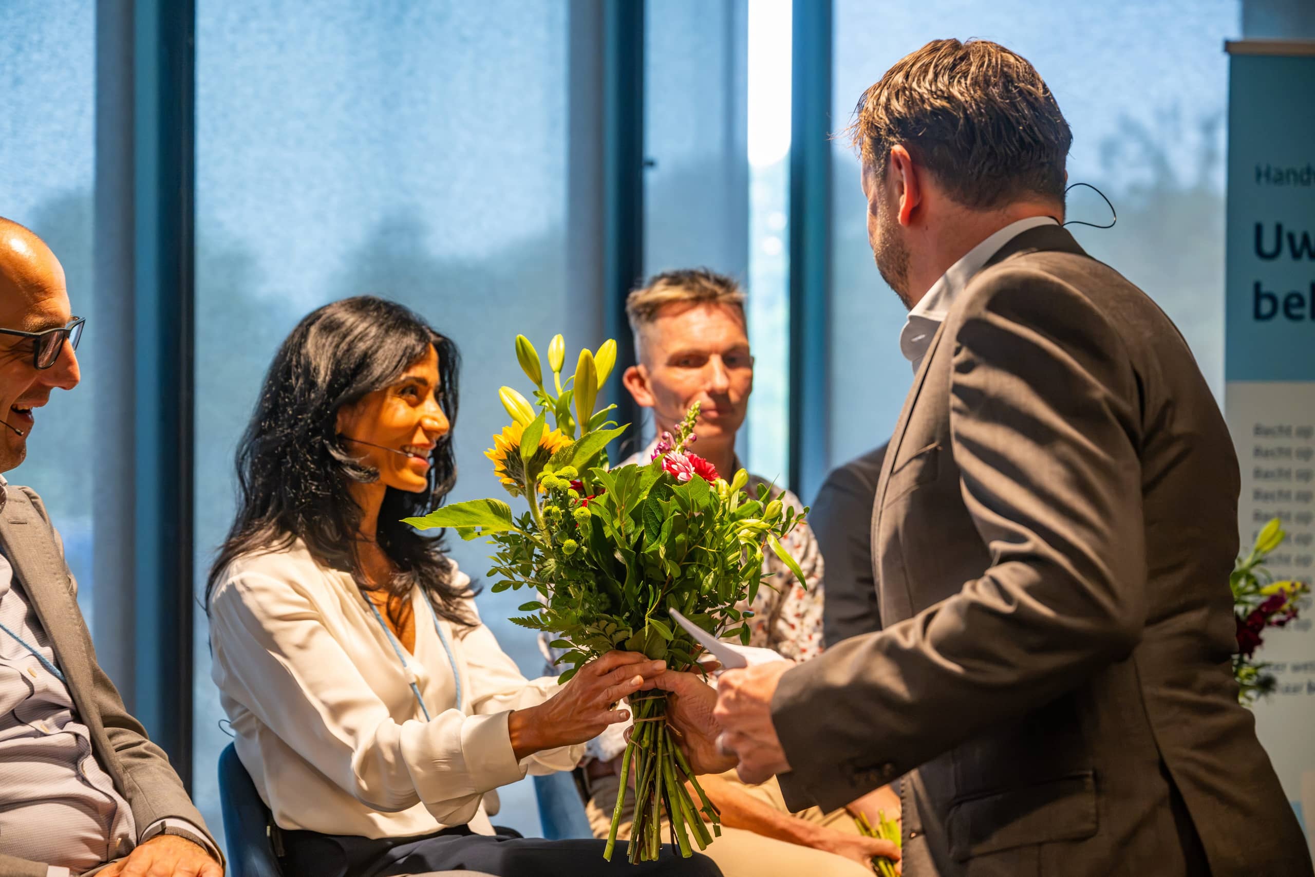 Hayde Zarkeshan glimlacht terwijl ze een grote bos bloemen in ontvangst neemt van een man in een grijs pak op het podium.